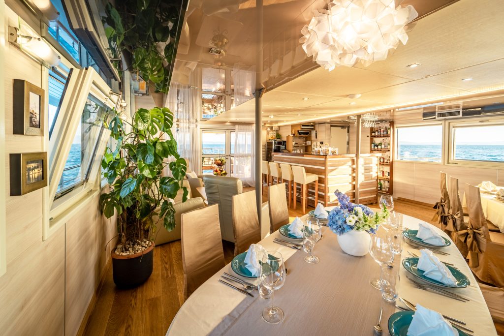 Elegant yacht interior with a dining table set for a meal, modern bar area, potted plant, and large windows showing ocean views. Warm lighting and stylish decor create a cozy, luxurious atmosphere.