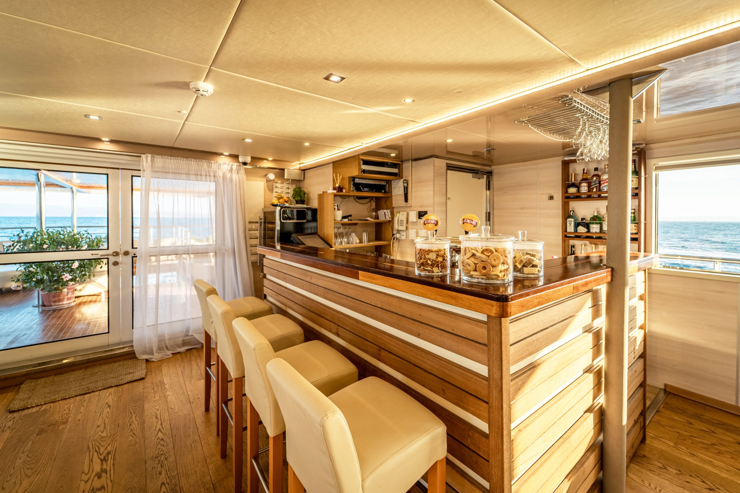 A bright, modern bar area on a boat with wooden floors and counters, cream bar stools, jars of cookies, shelves stocked with bottles, large windows, and ocean views in the background.