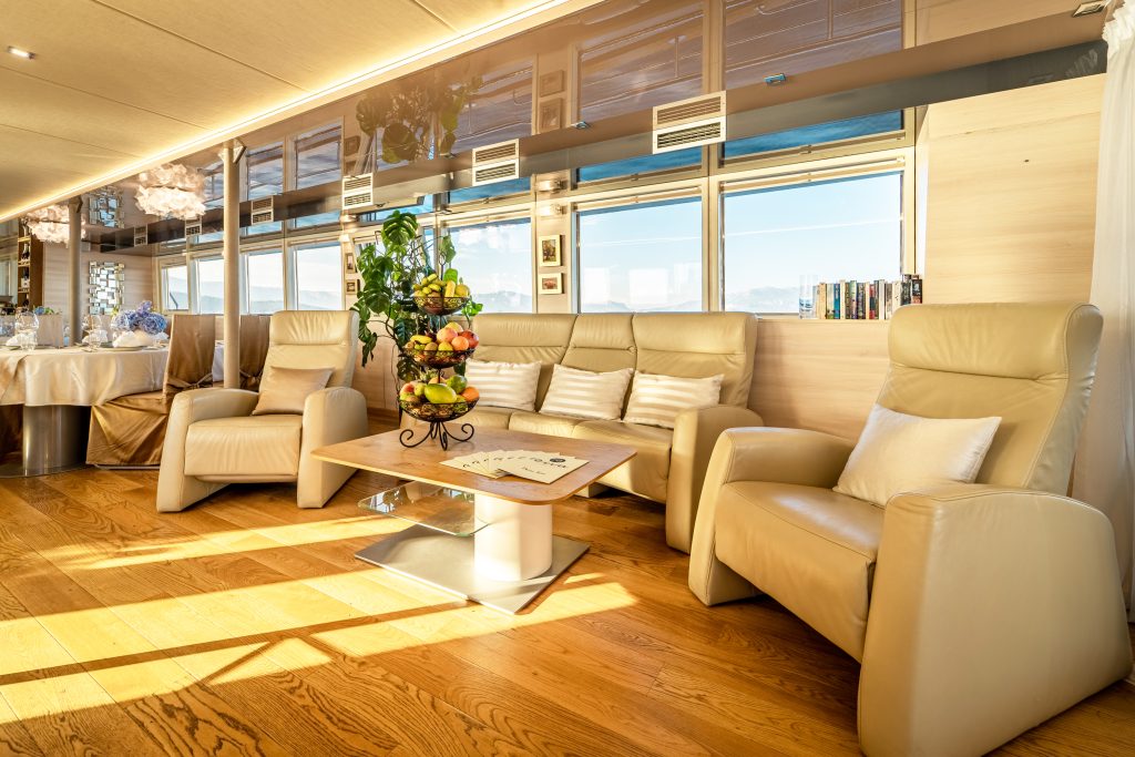 A bright, modern lounge with beige leather armchairs, a wooden floor, a table with a fruit centerpiece, large windows with blue sky views, and books on a shelf in the background.