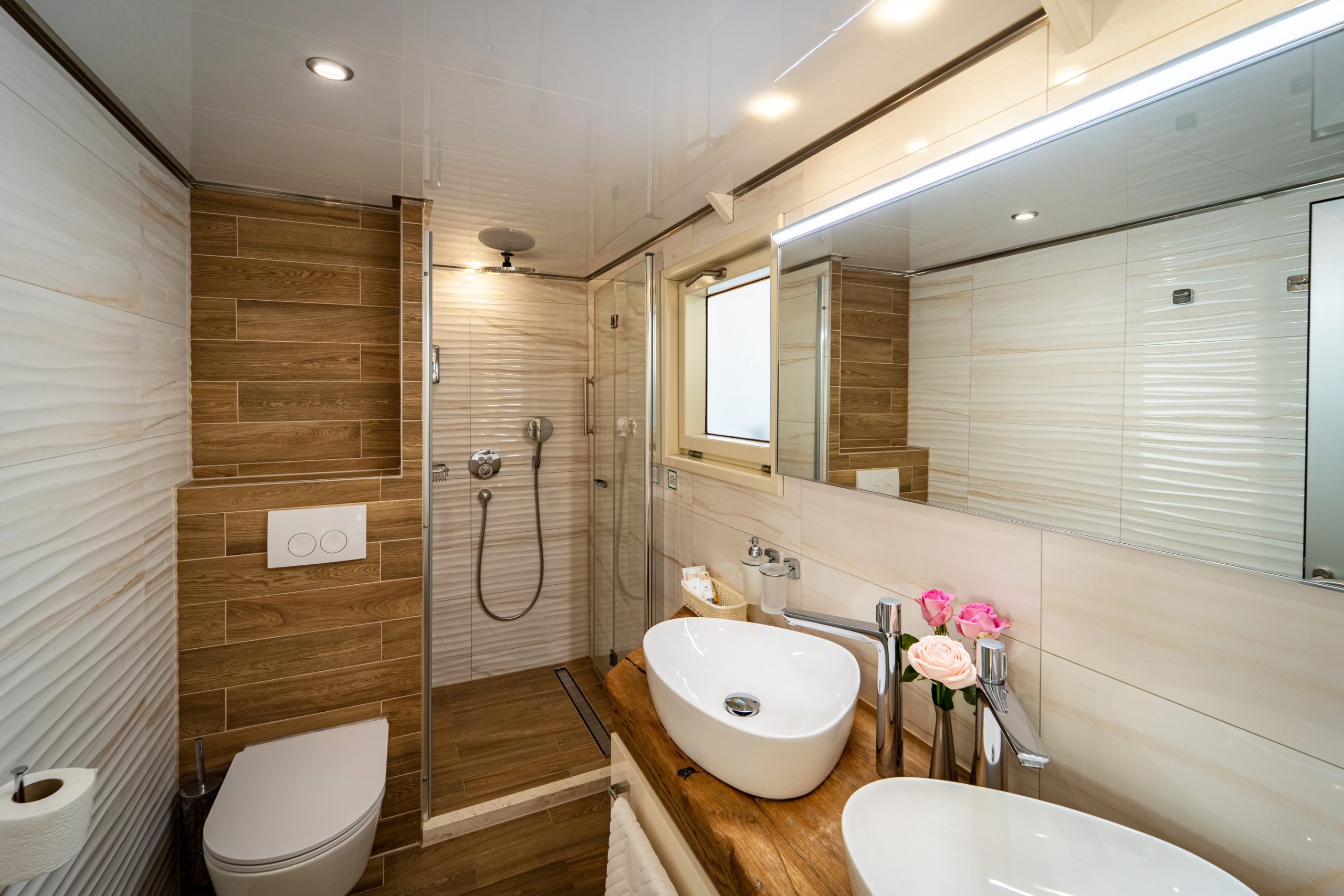Modern bathroom with wood-look tiles and white textured walls, featuring a walk-in shower, wall-mounted toilet, vessel sink on a wooden countertop, mirror, and a vase with pink flowers.