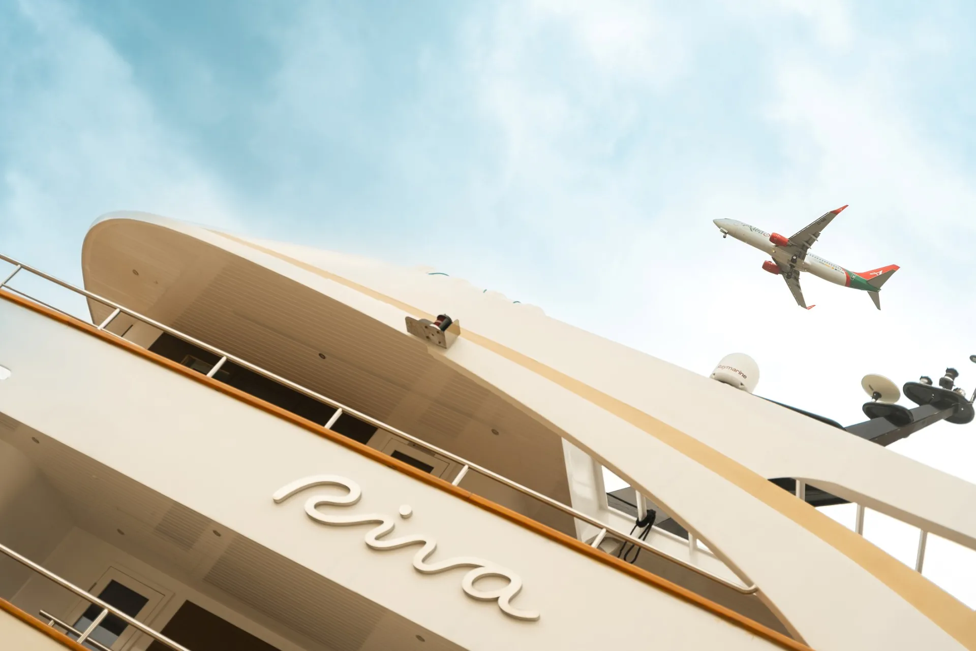 A commercial airplane flies above a white yacht named Riva under a blue sky with light clouds, viewed from a low angle.