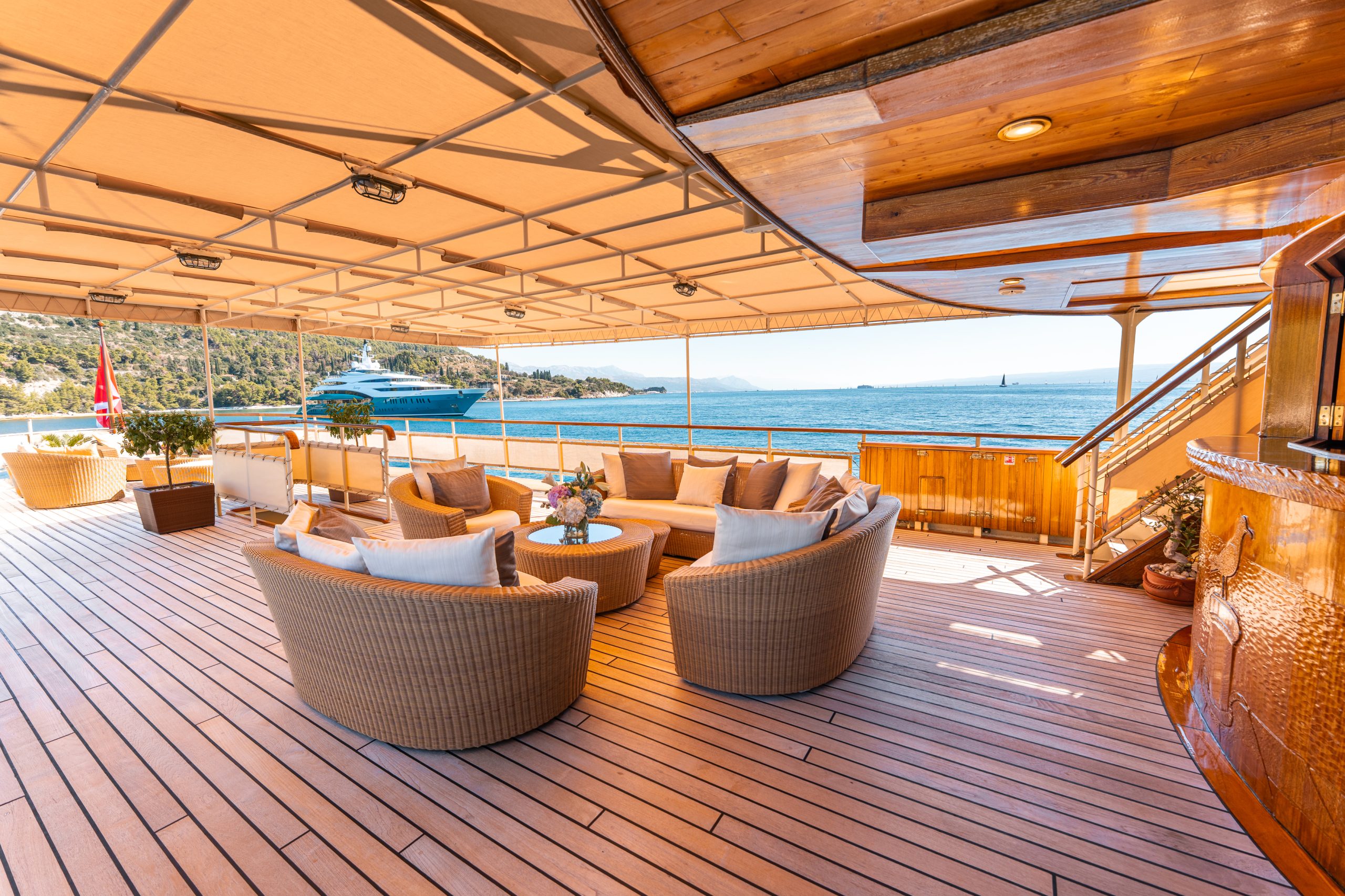 Spacious yacht deck with wicker seating, round tables, and potted plants, shaded by a canopy. Overlooks blue water, coastline, and another yacht in the distance under a bright sky.
