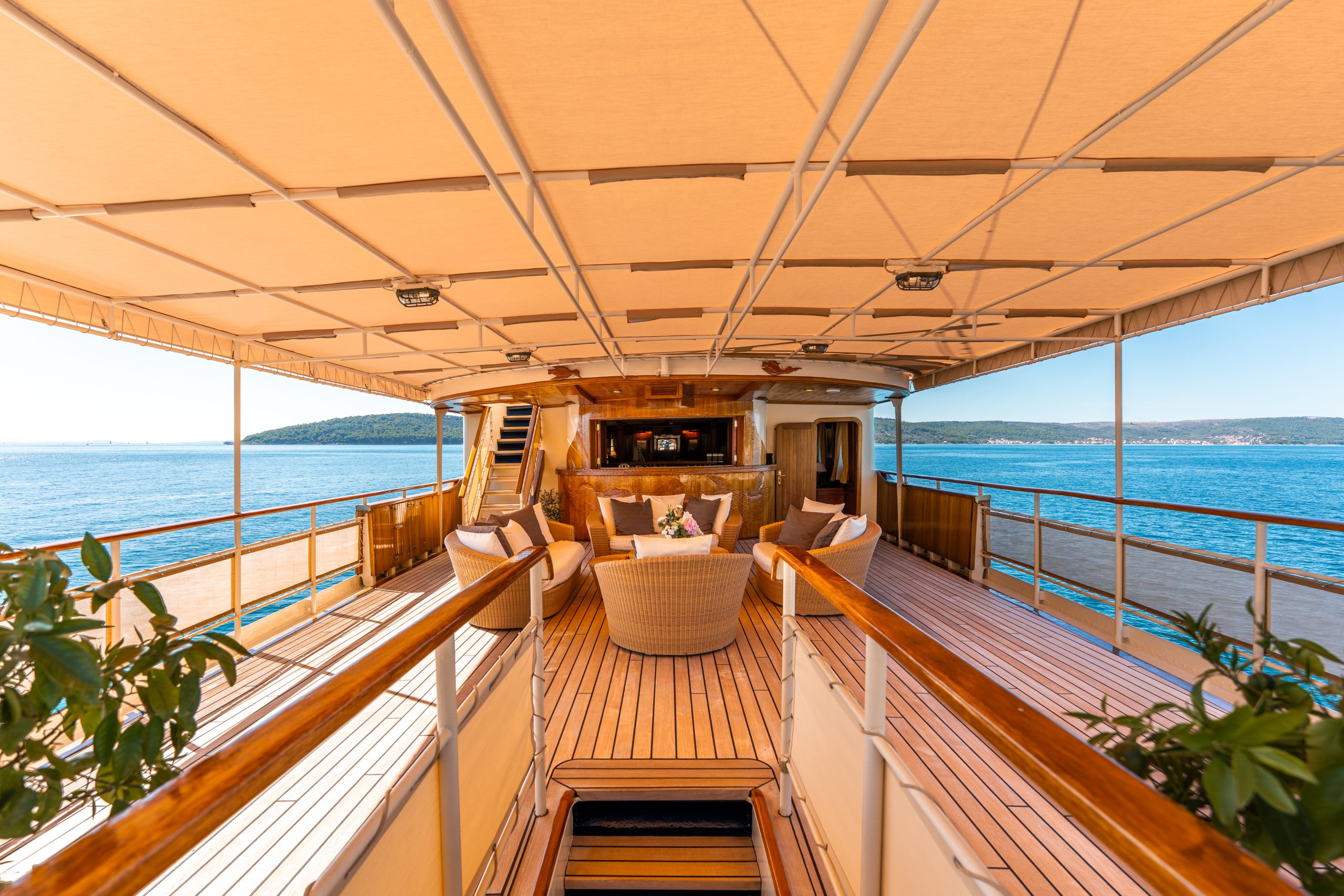 The deck of a luxury yacht with wooden flooring, wicker chairs around a table, potted plants, and a canopy overhead, overlooking calm blue water and distant hills under a clear sky.