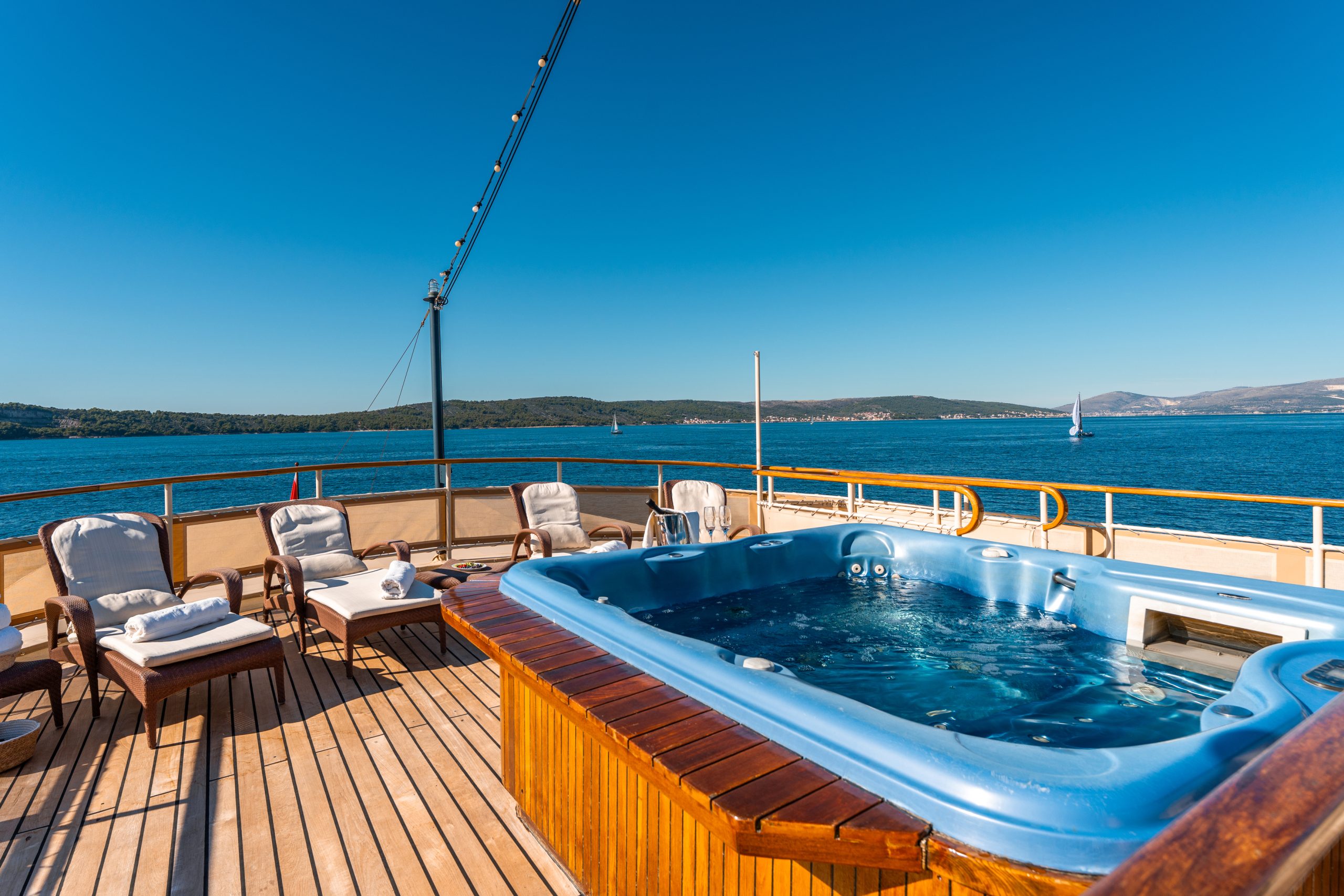 A luxurious yacht deck features a blue hot tub, cushioned lounge chairs with towels, and a view of calm blue water and distant hills under a clear, sunny sky.