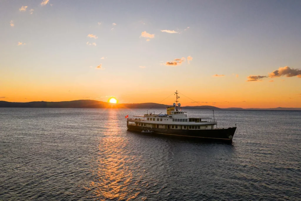 A large yacht floats on calm water at sunset, with the sun setting behind distant hills and a colorful sky reflected on the surface.