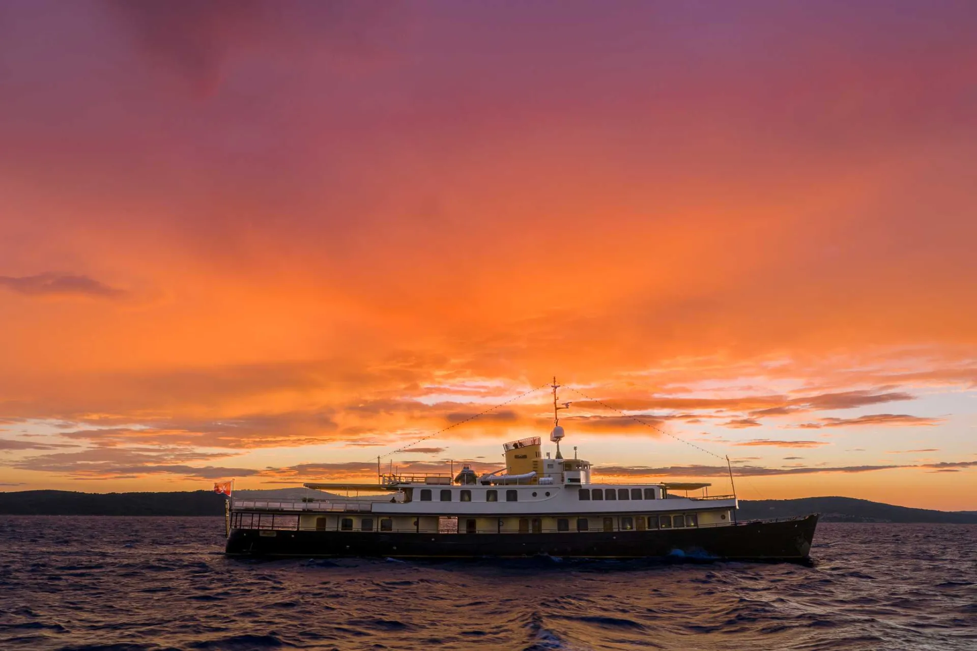 A ferry sails on the ocean under a vibrant orange and purple sunset sky, with gentle waves and distant hills on the horizon.