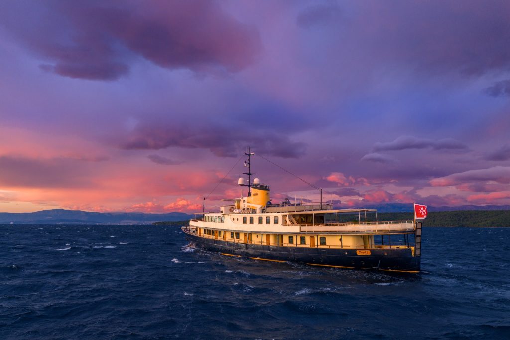 A vintage ferry boat sails on choppy blue water under a dramatic, colorful sunset sky with purple and pink clouds. A shoreline is visible in the distance.