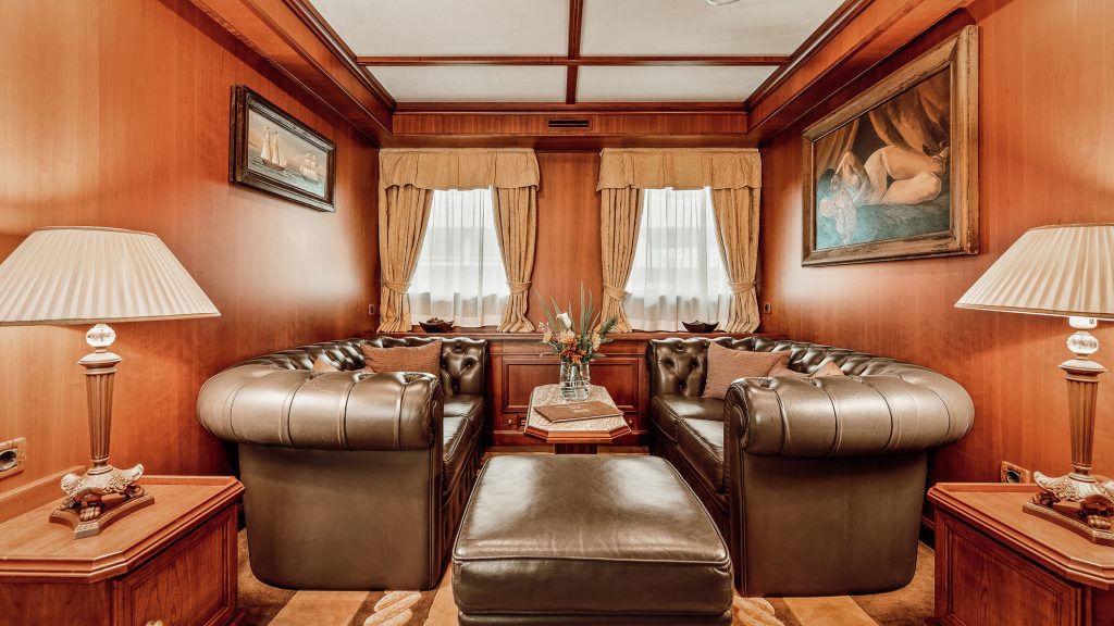 A cozy, classic sitting room with brown leather sofas, wood-paneled walls, two lamps, a central ottoman, curtains on two windows, and framed paintings on the walls. A flower arrangement is on a small table.