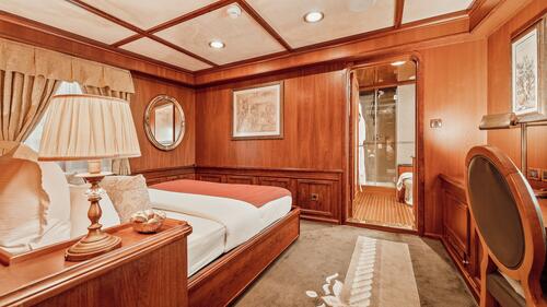 Luxurious wooden-paneled bedroom with a double bed, lamp, and mirror. An open door reveals an adjoining bathroom with a glass shower. Warm lighting creates a cozy and elegant atmosphere.