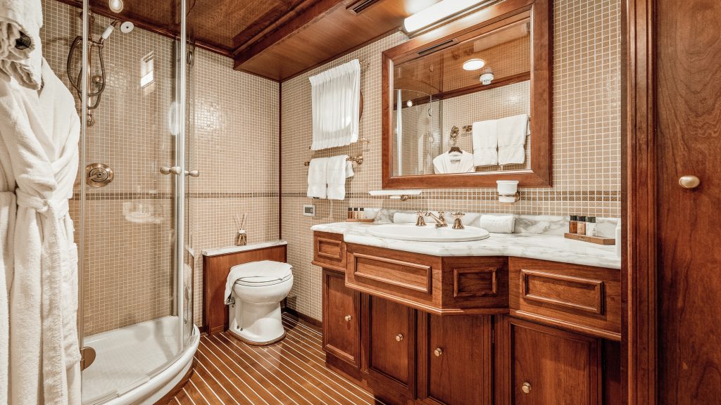 A cozy bathroom with wood-paneled cabinets, a marble countertop, a glass shower, a toilet, beige mosaic tile walls, and striped wooden flooring. White towels and robes are neatly hung.