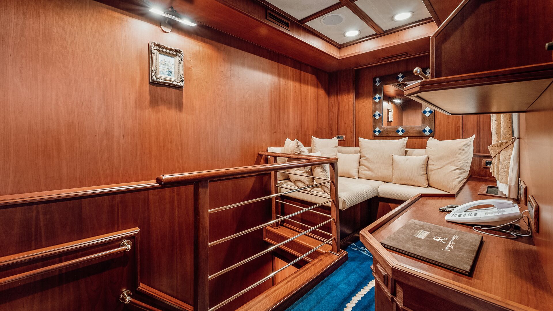 A cozy room with warm wooden walls, a white cushioned sofa, a small desk with a phone and folder, and a staircase with a metal railing. Blue accents appear in the carpet and a mirror with blue decorations.