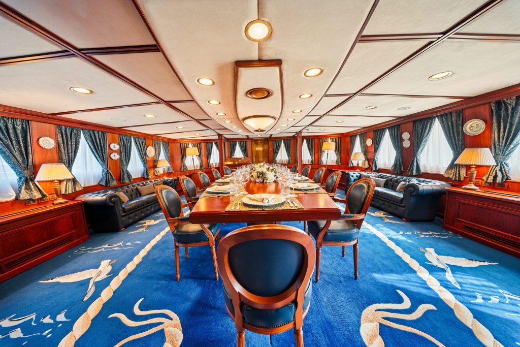 A luxurious dining room features a long table set for a meal, surrounded by black chairs, leather sofas, elegant curtains, wood paneling, and decorative lamps, with a blue carpet accented by white patterns.
