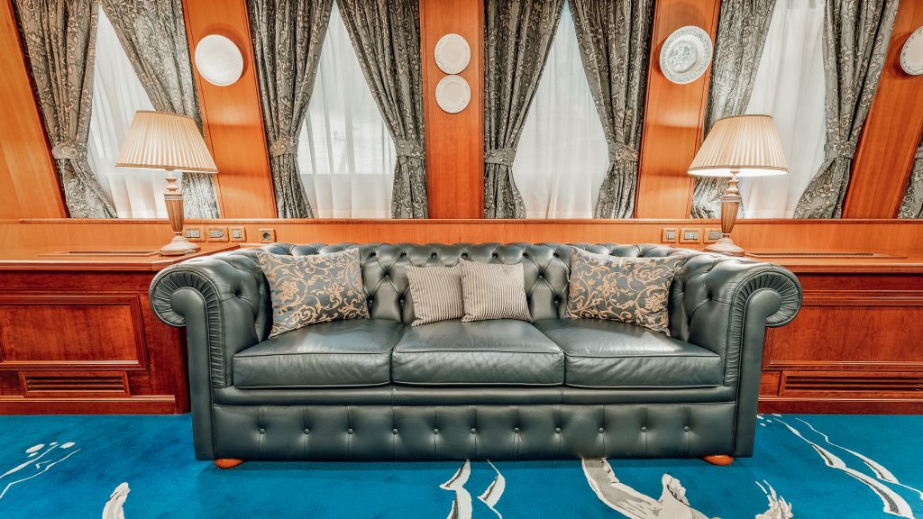 A luxurious green leather sofa with patterned and plain cushions sits on a blue carpet between two table lamps, against a wood-paneled wall with curtains and decorative plates.