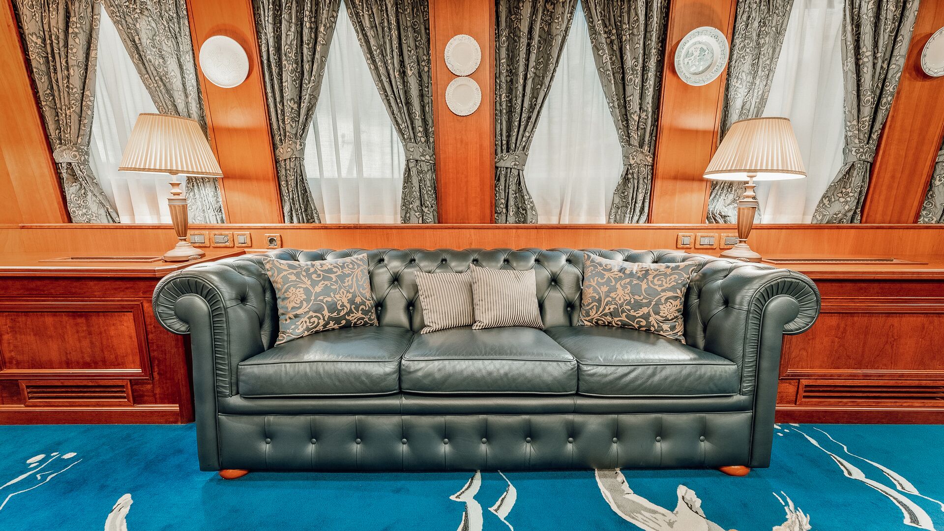 A luxurious green leather sofa with patterned and plain cushions sits on a blue carpet between two table lamps, against a wood-paneled wall with curtains and decorative plates.