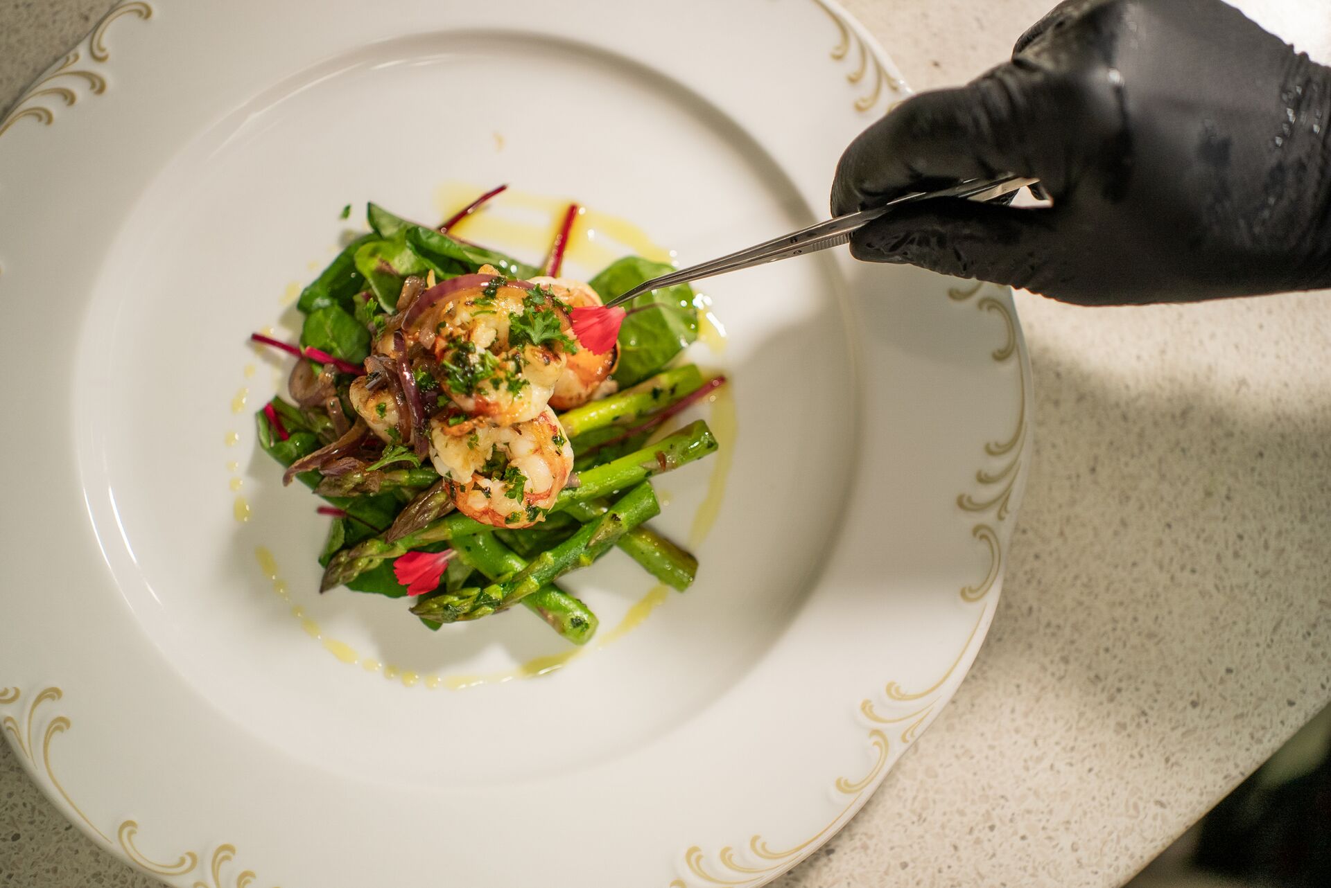 A gloved hand uses tongs to arrange shrimp atop a bed of asparagus and greens on a white decorative plate, garnished with herbs and drizzled with oil.