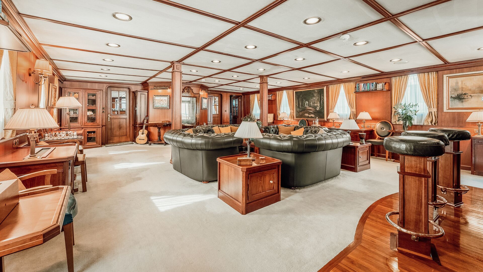 Luxurious yacht lounge with dark leather sofas, wood-paneled walls and furniture, lamps, bar stools, a guitar, and nautical decor. Large windows with curtains let in natural light.