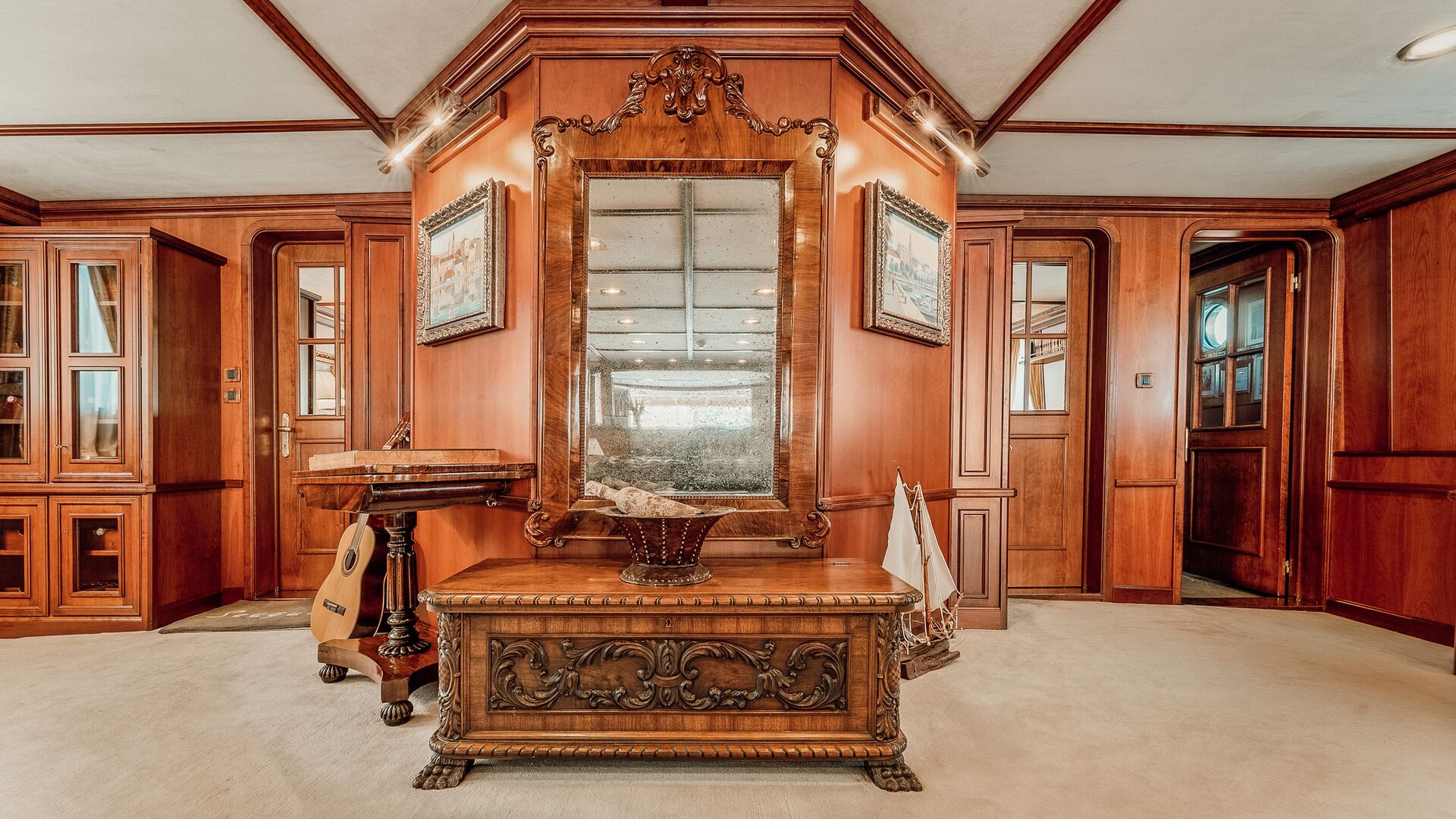 An ornate wooden bench with intricate carvings sits below a large, framed mirror on a wall with rich wood paneling. A decorative basket, small table, and model sailboat accent the elegant, classic interior.