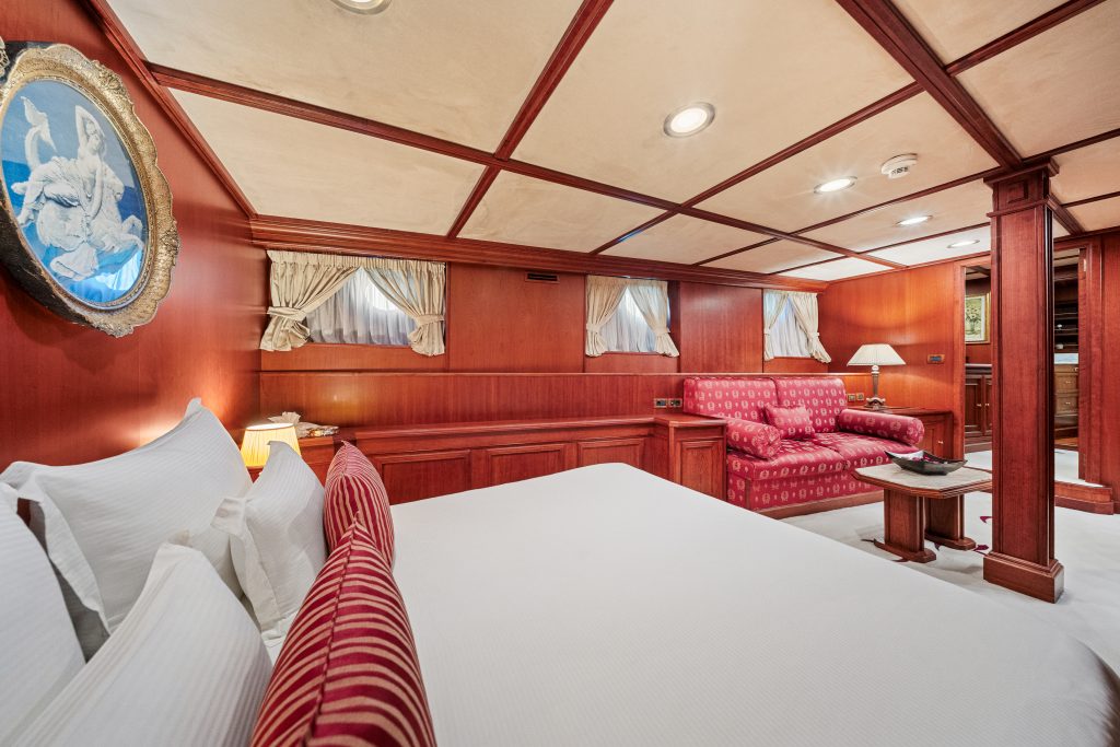 Luxurious cabin with wood-paneled walls, white bedding, and red-striped pillows. There is a red patterned sofa, small table, and framed artwork. Multiple windows with curtains let in natural light.