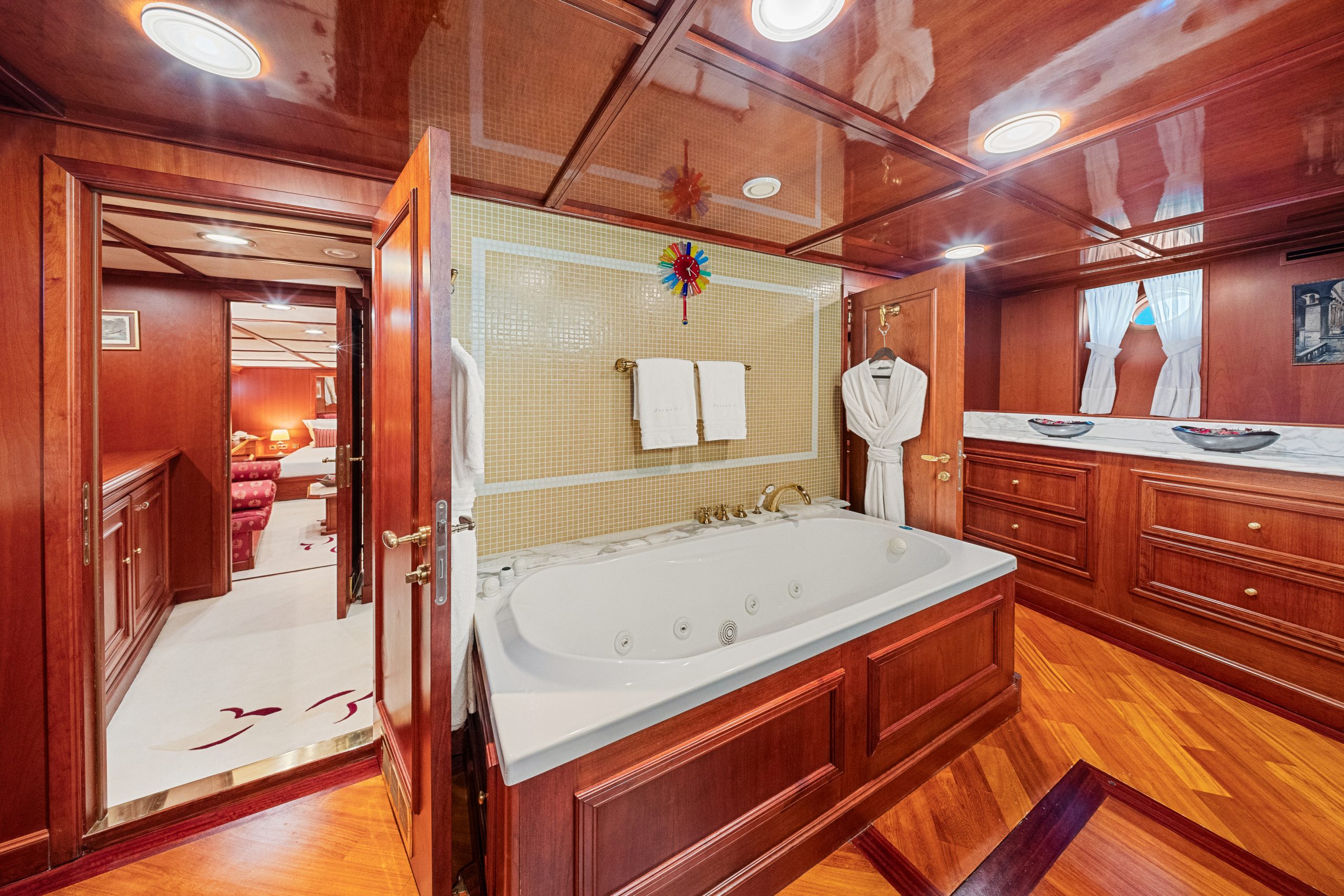 Luxurious bathroom with wooden walls and floors, a large white jacuzzi tub, gold fixtures, towels, and a robe. An open door reveals another room with red seating and white carpet.