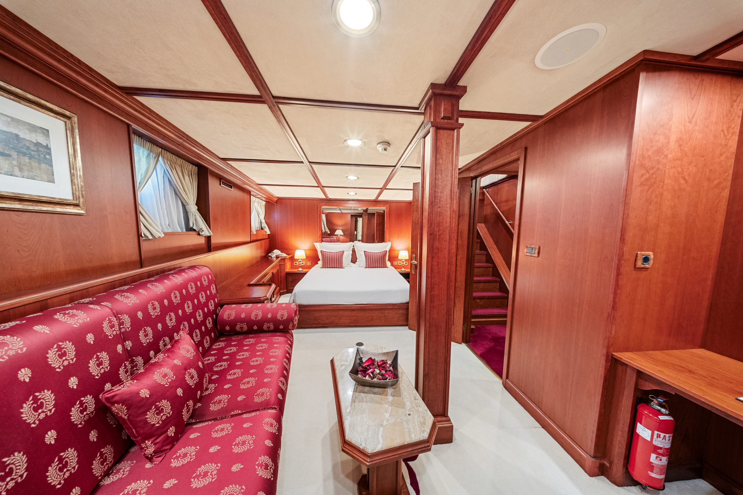 A cozy room with polished wood paneling, a bed with white linens and red cushions, a red patterned sofa, a small marble table, a staircase, and a fire extinguisher on the wall. Framed art and windows add decor.