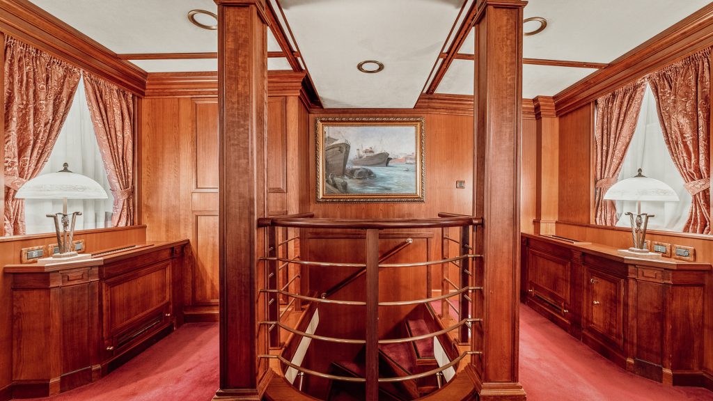 A luxurious hallway with rich wood paneling, red carpet, and gold railing around a staircase. Two ornate lamps sit on cabinets beneath curtained windows, and a framed seascape painting hangs on the wall.