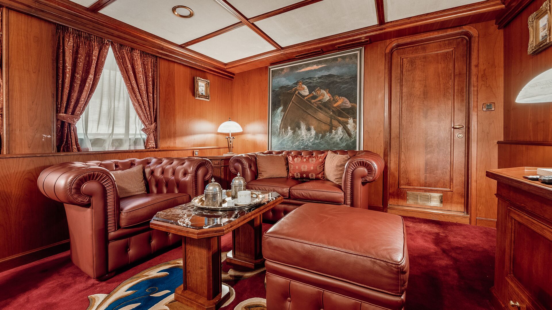 A luxurious room with rich wood paneling, maroon leather armchairs, a matching ottoman, a marble coffee table with a tea set, deep red carpet, and a painting of a lifeboat at sea on the wall.