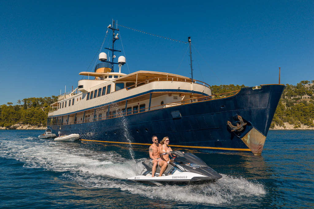 A man and woman ride a jet ski on blue water near a large luxury yacht, under a clear sky, with a forested shoreline in the background.