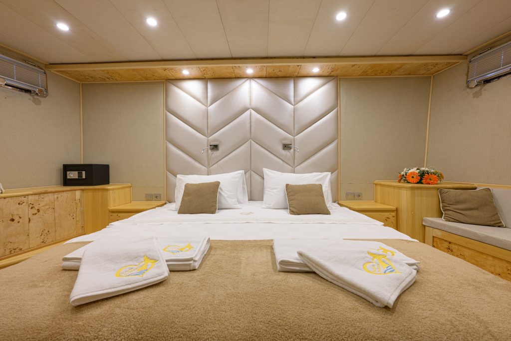A neatly made bed with white linens and beige accent pillows in a modern, well-lit room. Folded towels are placed on the bed, and there are built-in wooden cabinets and a small bouquet of flowers on the side.