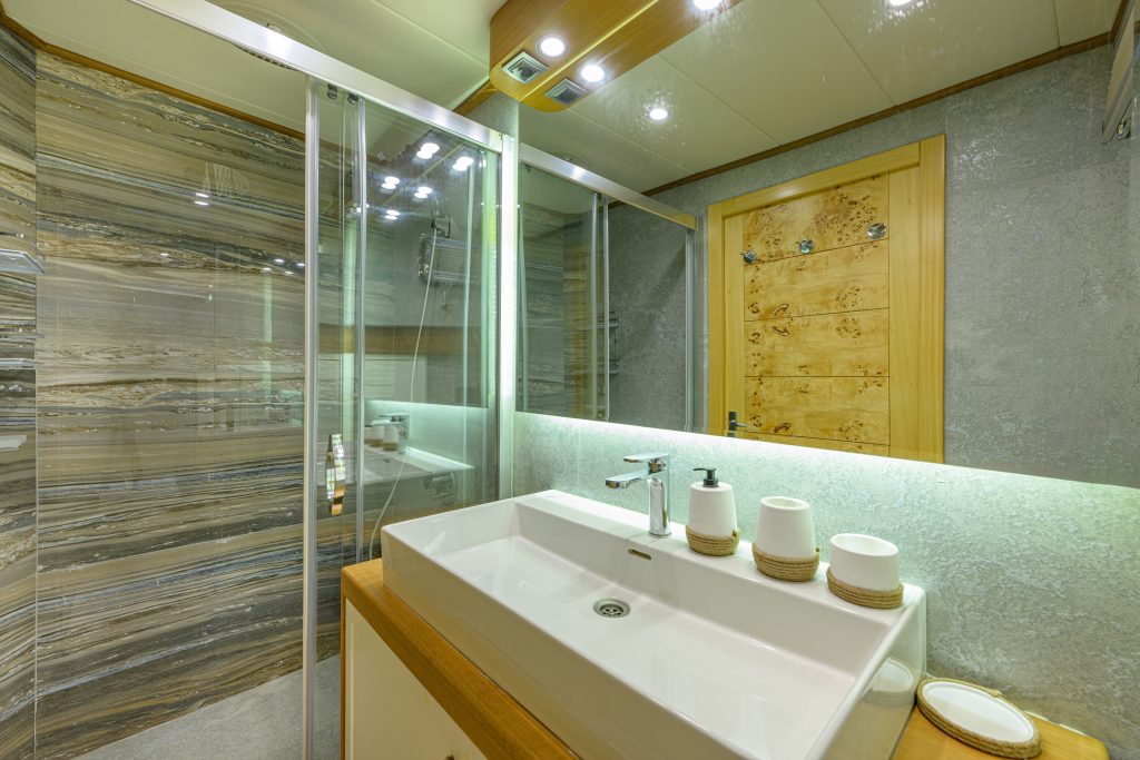 Modern bathroom with a large white rectangular sink, three white containers on the counter, a glass shower enclosure with marbled wall tiles, and a wooden door reflected in a large mirror.