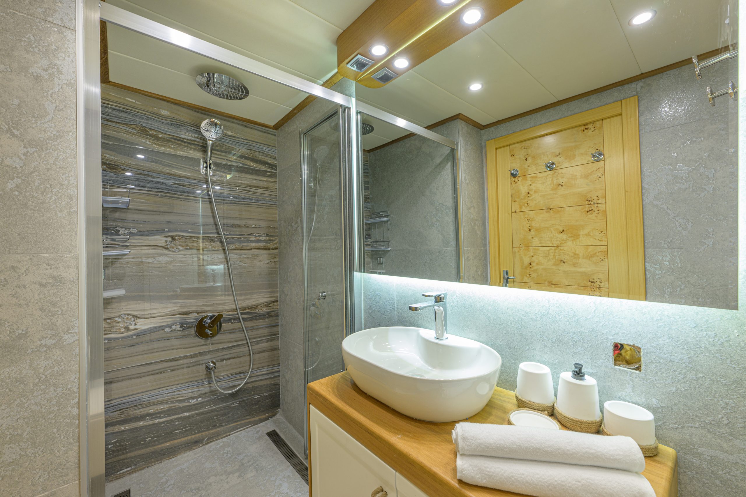 Modern bathroom with a glass-enclosed shower, rain showerhead, striped tile walls, vessel sink on a wooden countertop, rolled white towels, and a large mirror above the sink.