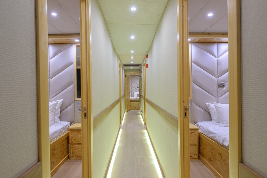 A brightly lit hallway with wooden floors and walls, lined with small rooms on each side. Each room has a single bed with white bedding and padded, light-colored headboards. Ceiling lights illuminate the corridor.