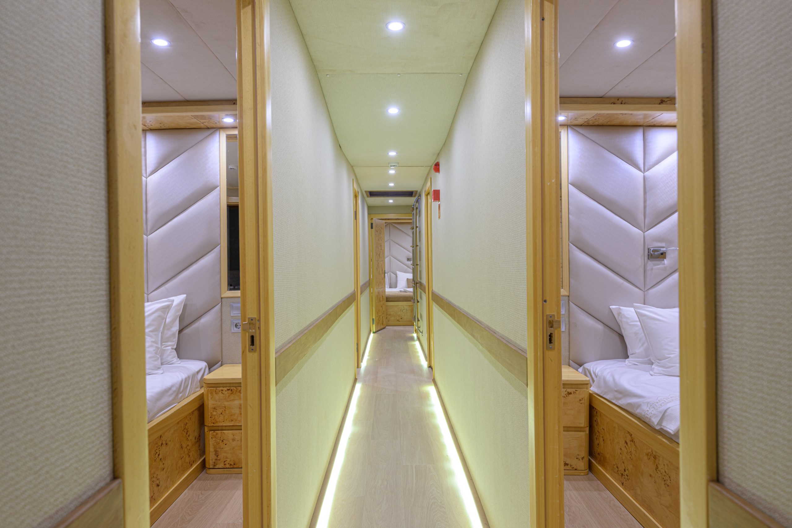 A brightly lit hallway with wooden floors and walls, lined with small rooms on each side. Each room has a single bed with white bedding and padded, light-colored headboards. Ceiling lights illuminate the corridor.