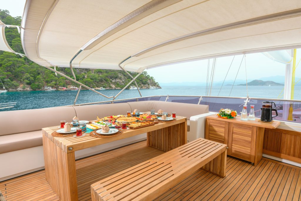 A wooden dining table set with breakfast food and drinks sits on the deck of a yacht, with benches and a view of the sea and distant green hills under a canopy.