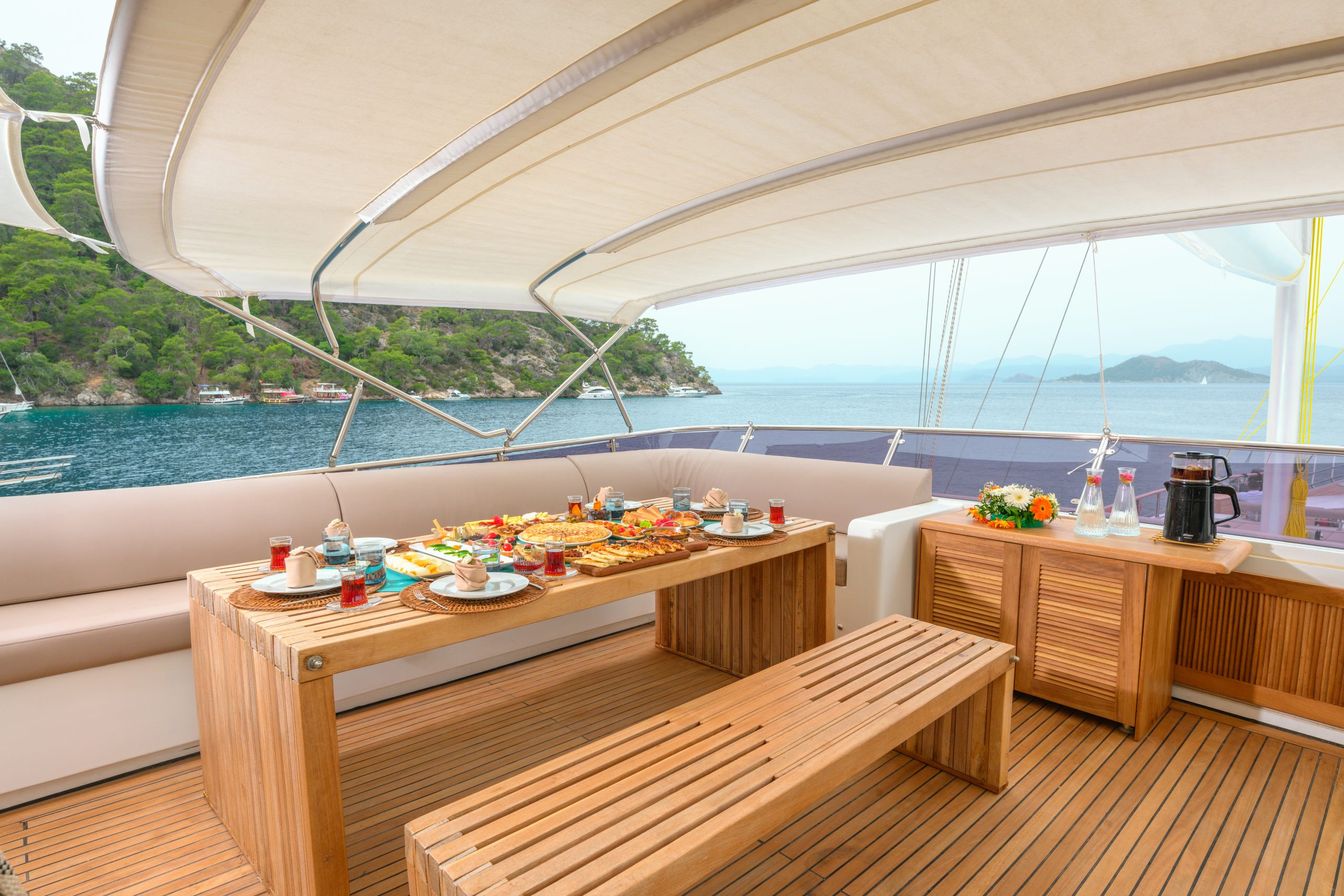 A wooden dining table set with breakfast food and drinks sits on the deck of a yacht, with benches and a view of the sea and distant green hills under a canopy.
