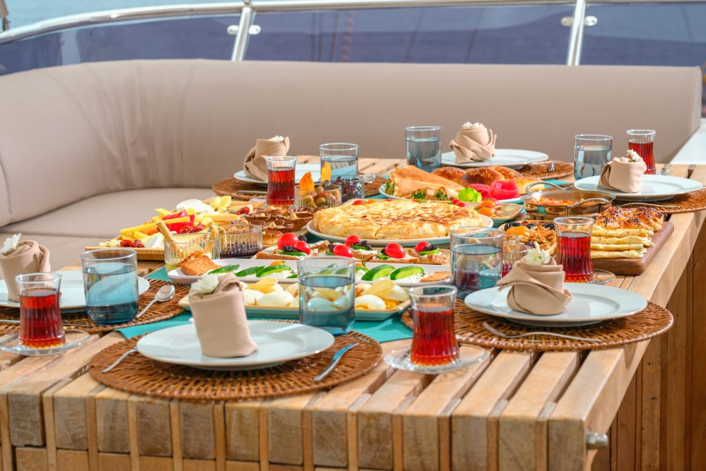 A breakfast table on a yacht with plates, glasses of tea, napkins, and a variety of foods including bread, fries, cheese, eggs, salad, and pastries, set on a wooden table with cushioned seating in the background.