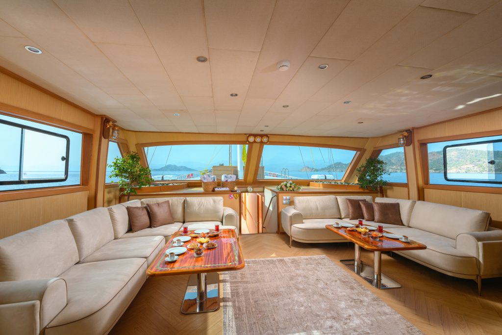Spacious yacht interior with large windows, beige sofas, wooden tables set with snacks and tea, potted plants, and a view of the sea and mountains outside. Natural light fills the room.