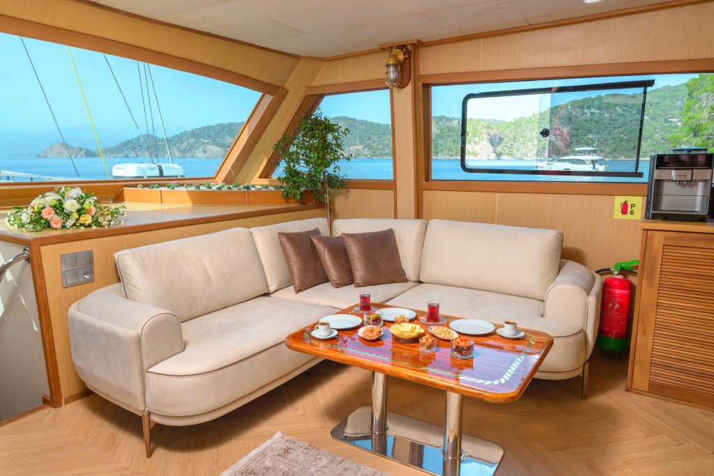 A cozy yacht interior with a curved beige sofa, a wooden table set for breakfast, large windows showing a scenic sea view, and accents like a potted plant, fire extinguisher, and coffee machine nearby.