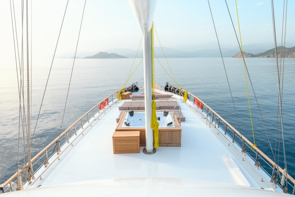 View from the deck of a yacht sailing on calm blue water, with distant mountains and clear sky in the background. The deck features seating areas and railings on both sides.