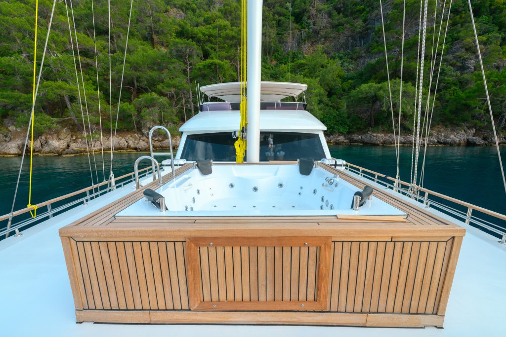 A luxury yacht features a built-in hot tub with wooden paneling on its deck, anchored near a lush, green forested shoreline. The calm water and trees are visible in the background.