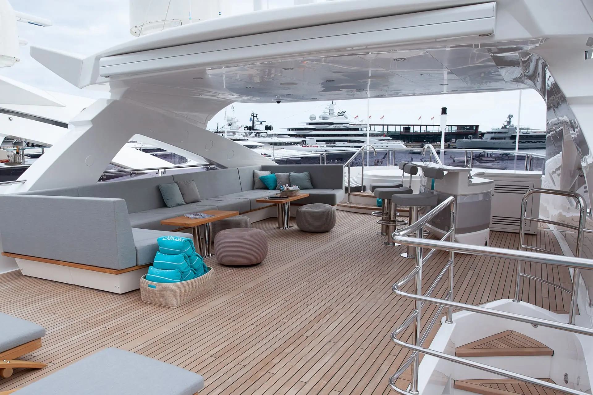 Luxury yacht deck with light gray cushioned seating, small tables, ottomans, and teal towels. Stainless steel railings and marina with other yachts are visible in the background under a cloudy sky.