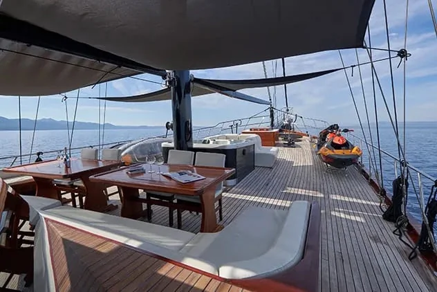 Luxurious yacht deck with wooden tables, cushioned seating, and a hot tub, overlooking calm blue water and distant mountains under a partly cloudy sky.