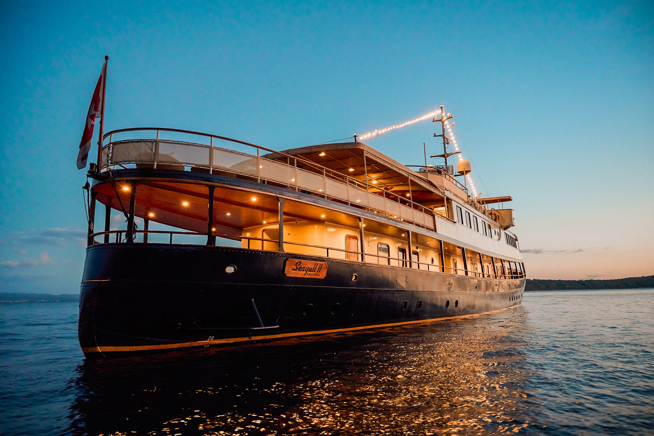 A large, elegant yacht with glowing lights is anchored on calm water at sunset, with a clear sky in the background and a flag flying at the stern.