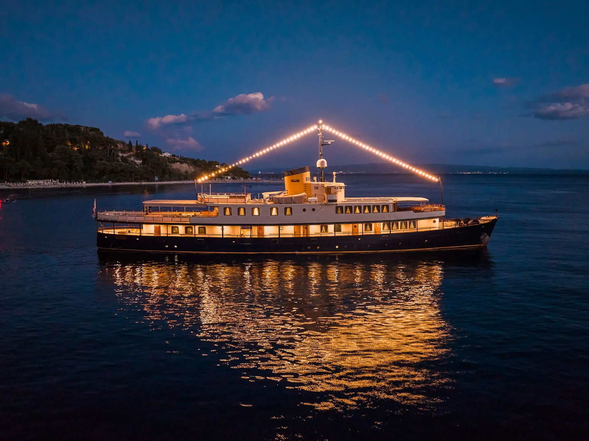 A large yacht decorated with string lights is anchored on calm water at dusk, reflecting its lights on the surface. The coastline with trees and buildings is visible in the background under a darkening sky.