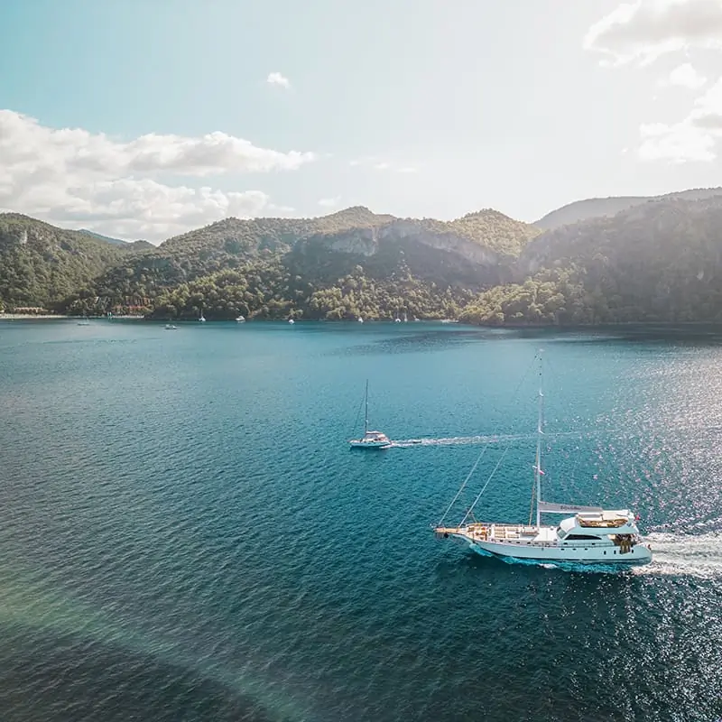 Two sailboats are sailing on calm blue water near a forested shoreline with hills in the background under a partly cloudy sky.