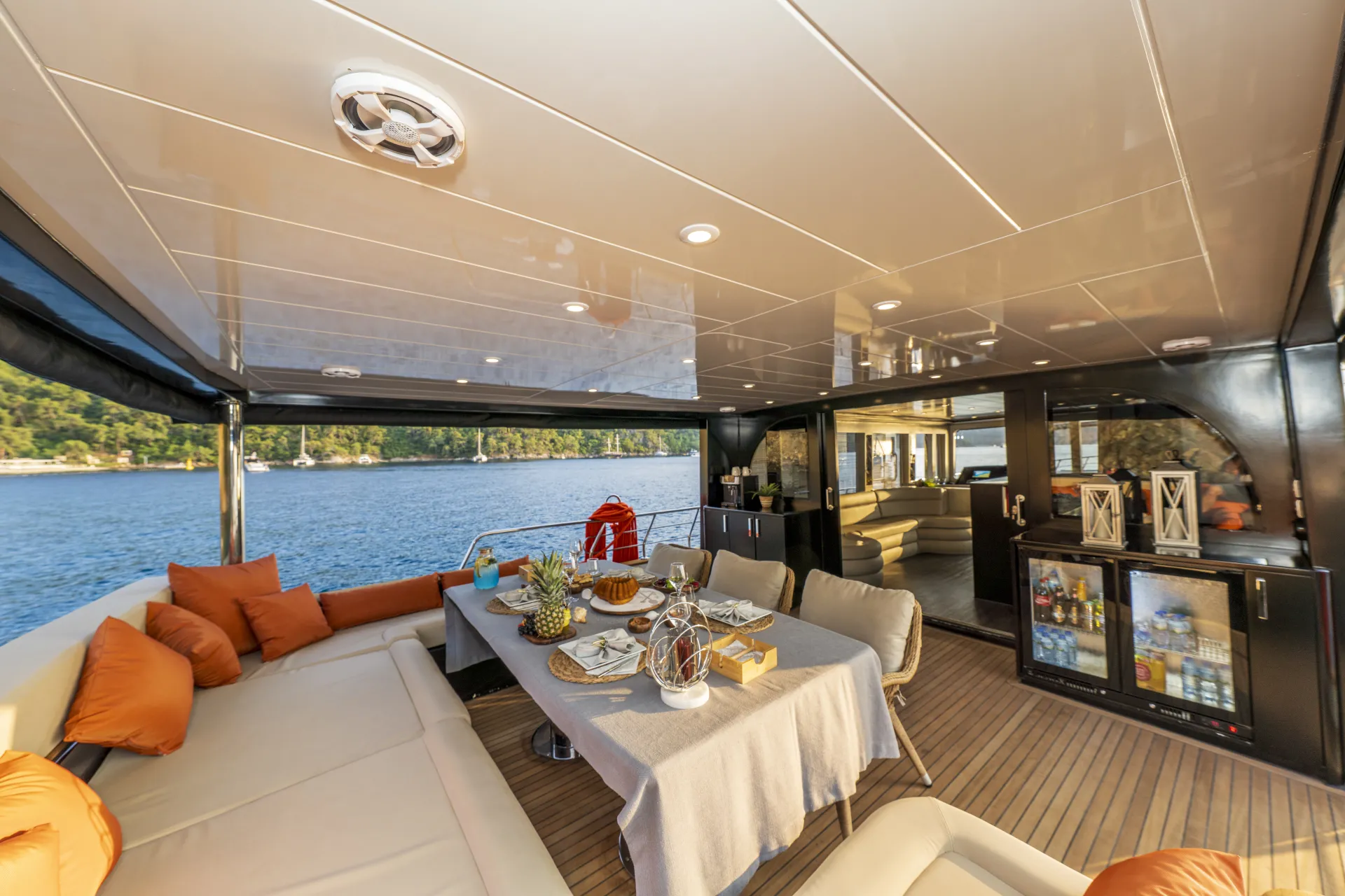 JUNIOR SIMAY A spacious yacht deck with cushioned seating, a dining table set with food and drinks, and a view of calm water and green shoreline in the background under a bright sky.