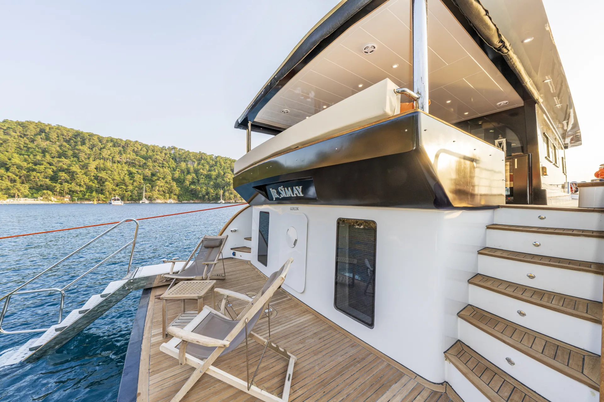 JUNIOR SIMAY A luxury yacht docked by a calm blue sea, featuring wooden deck chairs, steps leading to the upper deck, and a forested hillside in the background under clear skies.
