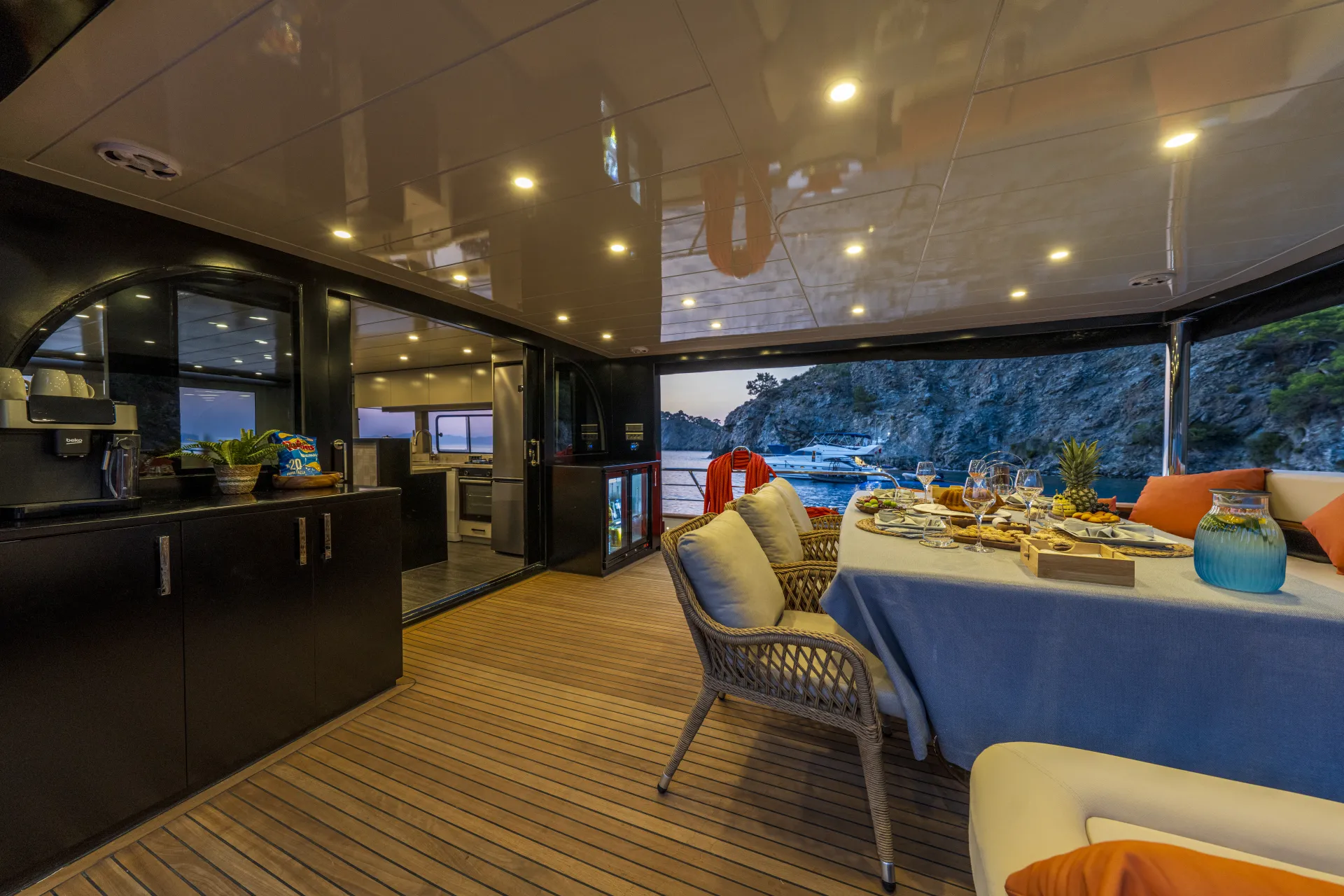 JUNIOR SIMAY Luxurious yacht deck at dusk with a set dining table, cushioned chairs, and ambient ceiling lights. The view overlooks calm water, nearby boats, and a rocky shoreline with trees in the background.