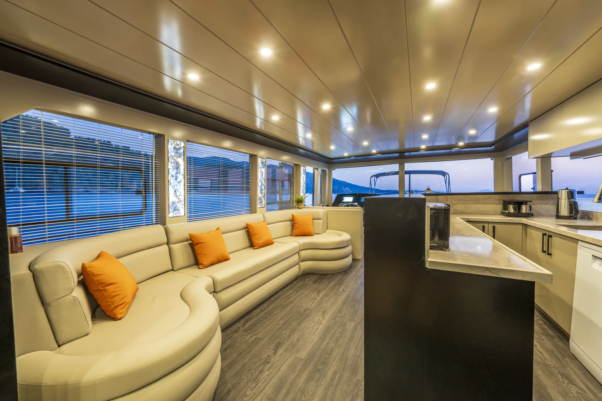 JUNIOR SIMAY Luxurious yacht interior with curved beige sofas adorned with orange pillows, large windows showing an ocean view at dusk, modern lighting, wood flooring, and a sleek kitchen area with countertops and appliances.