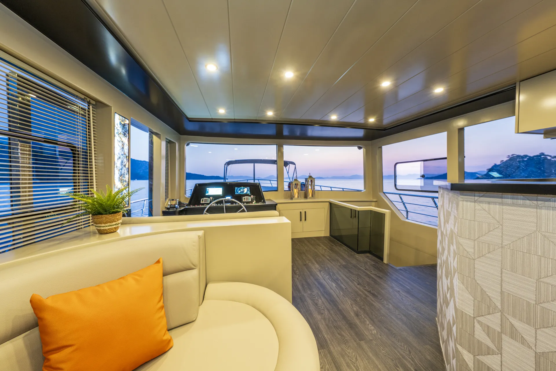 JUNIOR SIMAY Modern yacht interior with beige curved seating, an orange pillow, a potted plant, and large windows offering views of the sea at sunset. The spacious area features sleek decor and a helm station.