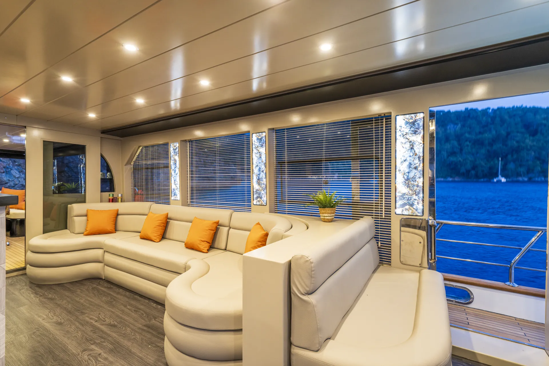 JUNIOR SIMAY Modern yacht interior with curved white leather sofas, orange throw pillows, potted plant, large windows with blinds, and view of blue water and green forested shoreline outside. Warm, recessed ceiling lights illuminate the space.